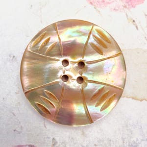 May include: A round, iridescent mother-of-pearl button featuring a four-hole center design. The surface displays a warm, golden-tan hue with subtle rainbow-like reflections and is decorated with four sets of three carved, linear grooves.