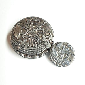 May include: Two circular silver-toned metal buttons featuring a relief design of a sailing ship on water. The larger button on the left and the smaller button on the right both display a mast, rigging, and a flag, with a textured background finish.
