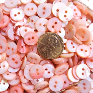 May include: A collection of small, round, pink buttons with two holes each. The buttons vary in shade from light to medium pink. A US penny is included for size reference.