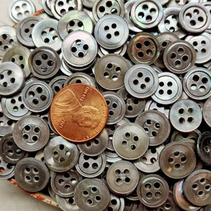 May include: A large collection of iridescent, silver-toned, four-hole buttons. The buttons are arranged in a random pattern and are all the same size. A US penny is included for scale.