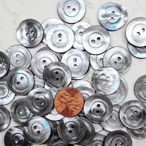 May include: A collection of approximately 30 black and iridescent mother of pearl buttons. The buttons are round and have two holes. The buttons are arranged in a random pattern.