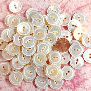 May include: A collection of white pearl buttons with two holes and a decorative design. The buttons are arranged in a pile on a pink surface.
