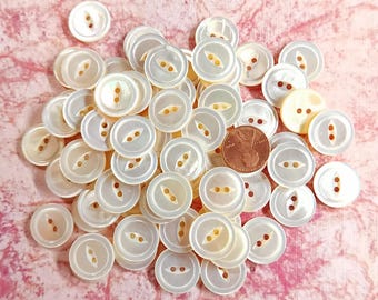 May include: A collection of white pearl buttons with two holes and a decorative design. The buttons are arranged in a pile on a pink surface.