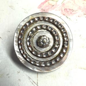 May include: A circular button featuring a central silver-toned flower motif surrounded by two concentric, textured metallic rings. The base of the button is composed of iridescent, off-white mother-of-pearl material, creating a layered, multi-dimensional appearance with a polished finish.