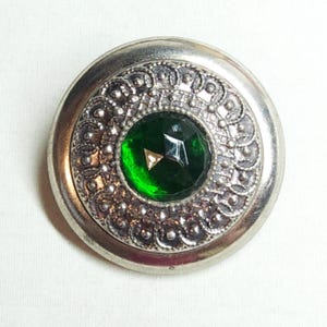 May include: A round, silver-toned button with an ornate, embossed design. The button features a large, faceted, emerald-green gemstone at its center. The button's design is symmetrical and detailed, suitable for sewing or crafting.