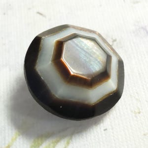 May include: A circular button featuring a multi-layered, faceted design. The center is a metallic, iridescent brown octagon, surrounded by a smooth white ring, and finished with a dark brown, faceted outer edge that slopes toward the base.