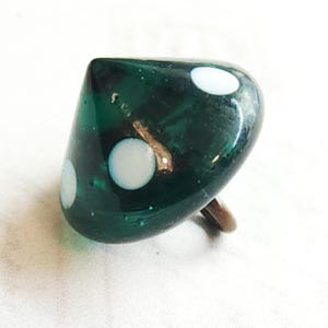 May include: A dark green glass dice-shaped ring with three white dots. The ring has a metal band and a rounded top. The dice is a unique accessory.