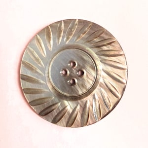 May include: A round, iridescent button with a smooth, recessed center featuring four holes. The outer edge is decorated with a radial pattern of elongated, leaf-shaped indentations that create a textured, sunburst effect across the surface.