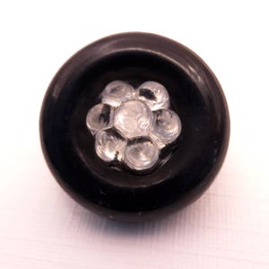 May include: A circular black button featuring a raised, clear, flower-shaped center element. The button has a smooth, glossy finish and a distinct outer rim, with the transparent, multi-petaled floral design set prominently in the middle of the dark base.