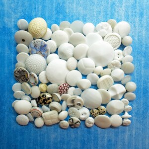 May include: A collection of various white buttons arranged in a square formation. The buttons feature diverse textures, including smooth, faceted, embossed, and patterned surfaces, with some displaying black, blue, or gold accents among the white.