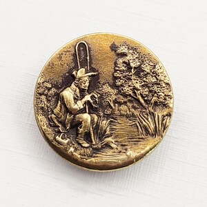 May include: A round, antique brass button with a detailed relief of a fisherman sitting by a river. The button features a man in a hat, holding a fishing rod, with trees and water in the background. The button has a vintage, aged appearance.