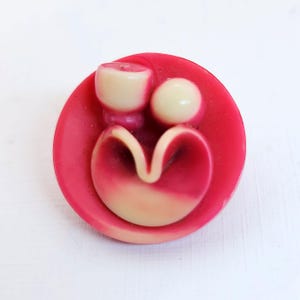 May include: A round, pink button with a unique design. It features a light beige, heart-shaped element at the bottom, a small sphere, and a cylindrical shape at the top. The button is likely made of plastic or resin.