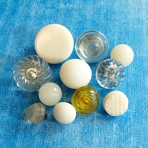 May include: A collection of ten assorted buttons in various shapes and materials. The set includes opaque white spherical buttons, clear glass buttons with swirl or textured patterns, a single translucent yellow button, and a small grey button.