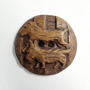 May include: A round, brown button featuring a textured surface with two central holes. The button is embossed with a detailed relief design of two dogs, one positioned above the other, with a wood-grain appearance throughout the entire piece.