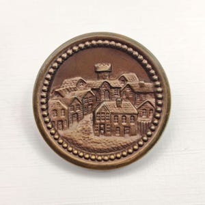 May include: A round, bronze-colored button with a detailed relief of a village scene. The button features small houses, a tower, and a dotted border. The design is raised, creating a textured surface.