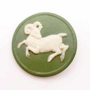 May include: A round, olive green cameo featuring a cream-colored ram in profile. The ram has detailed horns and is set against a solid green background. The cameo is a classic design, suitable for jewelry or decorative purposes.