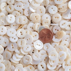 May include: A large collection of white mother of pearl buttons. The buttons are all round and have two holes. There is a penny in the image for scale.