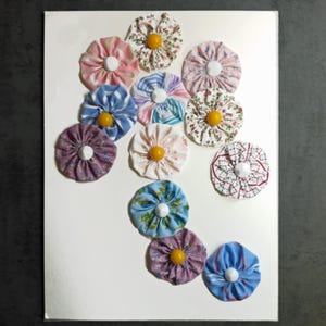 May include: A collection of twelve circular fabric yo-yos arranged on a white surface. Each piece features gathered fabric in various patterns including floral, striped, and plaid designs, with a central button in either white or yellow.