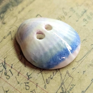May include: A handmade, oval-shaped button with two holes. The button features a blend of colors, including white, light purple, and blue. The blue color is concentrated on the bottom edge. The button is likely made of ceramic or a similar material.