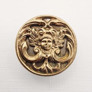 May include: A round, antique-style button with a detailed relief design. The button features a central cherub face with wings, surrounded by ornate scrollwork and a textured border. The color is a warm, aged gold.
