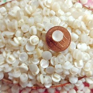 May include: A close-up image of a large pile of white, round, plastic buttons. A US penny is visible in the center of the image.