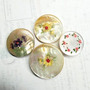 May include: Four round, mother-of-pearl buttons with hand-painted floral designs. The buttons feature yellow, purple, and pink flowers with green leaves. The buttons vary in size and design, with a vintage aesthetic.