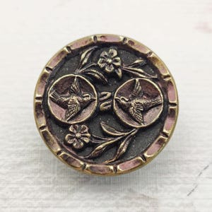 May include: Round, antique-style button with a bronze finish. The button features two circular medallions with bird designs, connected by a decorative element. Floral and leaf motifs surround the medallions. The button has a raised, textured edge.