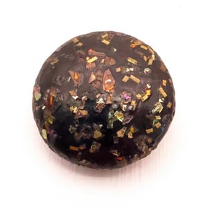 May include: A round, dark brown button with a smooth, glossy surface embedded with numerous small, reflective, multi-colored metallic flakes that catch the light, creating a speckled appearance across the entire circular face of the object.