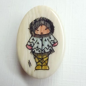 May include: Oval-shaped pendant with a hand-painted illustration of a person in winter clothing. The figure wears a hooded coat with a geometric pattern, yellow boots, and a fur-lined hood. The pendant is off-white with a slightly textured surface.