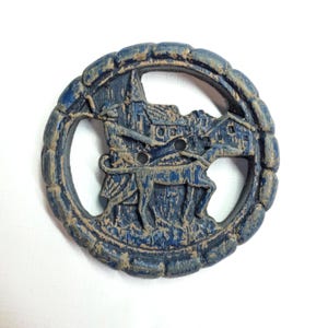 May include: A circular button with a distressed navy blue finish and tan highlights. The design features a relief carving of a person walking a dog in front of a building. The button has a scalloped border and two central holes for attachment.