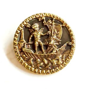 May include: A round button featuring a gold-toned metallic relief of a figure standing on a boat with a sail, set against a dark brown background. The edge of the button has a decorative, textured border with a repeating pattern of small indentations.