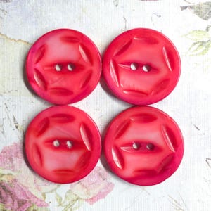 May include: Four circular, bright red buttons arranged in a two-by-two grid. Each button features a smooth, glossy finish with a central horizontal bar containing two thread holes and a series of curved, recessed indentations around the perimeter.