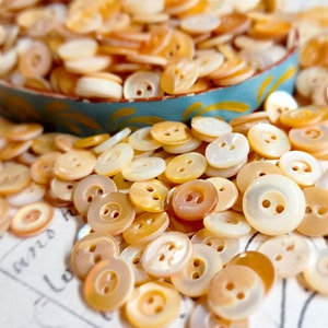 May include: A collection of various sizes of pearlescent buttons in shades of cream, tan, and gold. The buttons are scattered on a surface and in a small, turquoise and yellow painted bowl. The buttons have two or four holes for sewing.