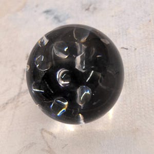 May include: A spherical glass object with a dark, translucent black base featuring embedded clear, bubble-like inclusions throughout the surface. The orb has a smooth, polished finish that reflects light, creating bright highlights on the clear spots.