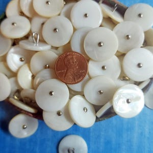 May include: A collection of white, round buttons with a pearlescent sheen and a silver-colored center. A copper penny is included for size reference. The buttons are scattered on a blue surface.