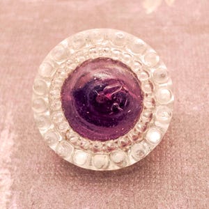 May include: A circular button featuring a deep purple, smooth, domed center surrounded by a clear, textured border composed of small, raised, bead-like glass elements arranged in a concentric pattern around the central purple core.