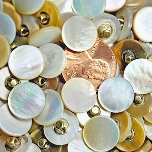 May include: A large collection of round, white, iridescent mother-of-pearl buttons with gold metal shanks. The buttons are arranged in a random pattern and are various sizes. A US penny is included for scale.