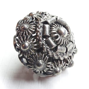 May include: A spherical silver ring featuring an intricate openwork design composed of multiple small flower-shaped motifs. The surface displays a textured, oxidized finish with a central circular element surrounded by a braided border detail.
