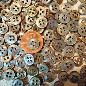 May include: A large pile of iridescent, mother-of-pearl buttons with four holes each. The buttons are a mix of light and dark gray colors. A US penny is placed on top of the buttons for scale.