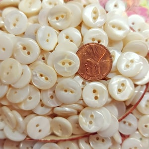 May include: A collection of off-white, pearlescent buttons with two holes each. The buttons are round and vary slightly in size. A U.S. penny is included for size comparison. The buttons are scattered, suggesting a craft or sewing supply.