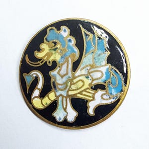 May include: A round, enameled pin featuring a stylized dragon design. The dragon is depicted in shades of blue, yellow, and white against a black background. The pin has a gold-colored border, adding a touch of elegance to the design.