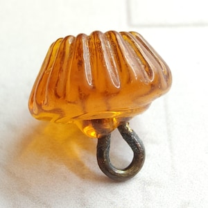May include: Amber-colored glass button with a ribbed, rounded top and a metal loop for sewing. The button has a vintage aesthetic, with a warm, translucent appearance. The button is a small, decorative element.