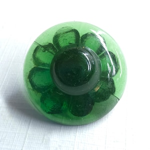 May include: A round, translucent green glass knob featuring a molded floral pattern with multiple petals radiating from a smooth, raised circular center. The glass has a glossy finish and a deep emerald hue, showing internal texture and light refraction.