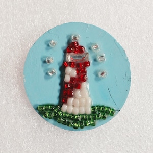 May include: A circular light blue disc featuring a beaded lighthouse design. The lighthouse is constructed from red and white beads, with green beads at the base representing grass and clear beads scattered around to represent the sky.