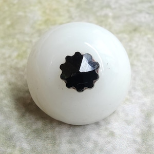 May include: A white, round decorative knob with a black, star-shaped center. The knob has a glossy finish and is likely made of glass or ceramic. This decorative knob is suitable for furniture or cabinet doors.