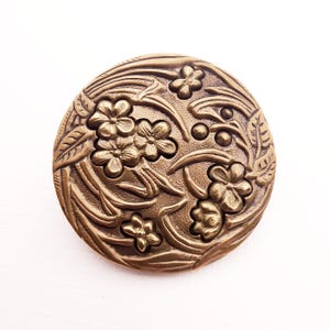 May include: A round, antique brass-colored button with a floral design. The button features embossed flowers, leaves, and vines. The button is set against a white background.