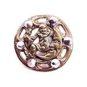 May include: A vintage brass button with a detailed design of a seated jester on a crescent moon. The button is adorned with six silver-colored, faceted embellishments around the edge. The jester has a ruffled collar and is holding a small object.