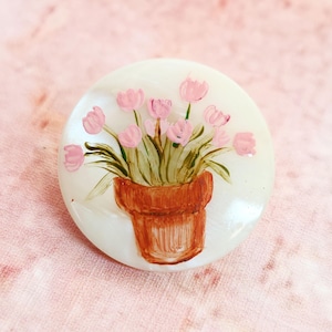 May include: A round, off-white brooch featuring a hand-painted design of pink tulips in a brown terracotta pot. The brooch has a glossy finish and is set against a soft pink background. The floral design adds a touch of spring.