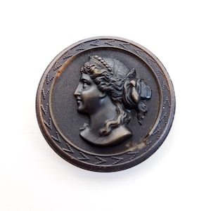 May include: A round, dark bronze-colored button with a detailed relief of a woman's profile. The woman has flowing hair and wears a decorative headpiece. The button has a textured border with a repeating pattern.