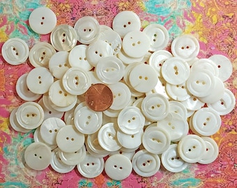 May include: A pile of white mother of pearl buttons with two holes each. The buttons are arranged in a random pattern and are all the same size. There is a US penny in the center of the pile for scale.